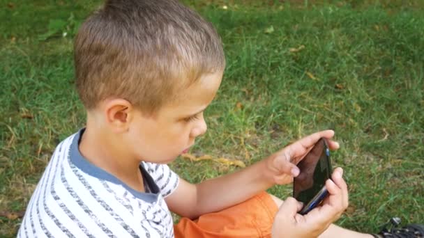 7歳の少年がスマートフォンでゲームをプレイします 子供が夏に公園の芝生の上に座っている 子供とスマートフォン 子供のための電子ゲーム — ストック動画