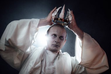 Ölü bir adamın makyajındaki bir adam kafasında kafatasları olan bir taç takar. Cadılar Bayramı için kasvetli gotik bir görüntü. Stüdyoda duman ve ışıkla yaratıcı fotoğrafçılık. Beyaz cüppeli bir şeytan.