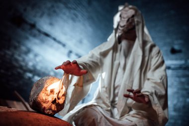 Veba doktoru. Cadılar Bayramı için kasvetli bir görüntünün kostüm oyunu. Kuş gagalı ve beyaz kapüşonlu bir maske. Maskeli bir adam ellerini yanan gözleriyle bir insan kafatasına doğru uzatıyor.