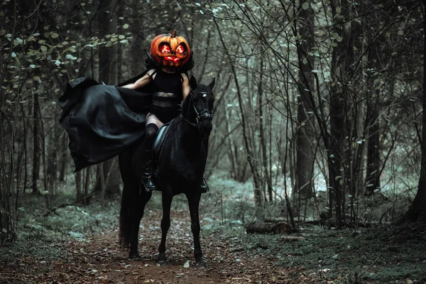 骑手正骑在马背上 一个穿着黑色长雨衣 戴着南瓜面具 眼睛灼热的女孩 在秋天的森林里 鬼鬼祟祟地为万圣节拍摄科幻片 风中飘扬的斗篷 — 图库照片