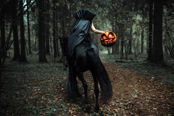 形状为南瓜 眼睛灼热 令人毛骨悚然的女孩服装之间 穿着黑色长斗篷的骑手牵着马的缰绳 秋天森林里一个阴郁形象的滑稽表演 — 图库照片