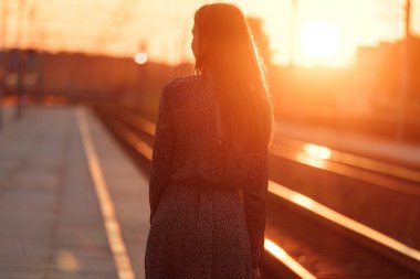 Tren peronu gün batımında. Gün batımında güneşin güzel altın ışığı. Trenle yolculuk zamanı. Kız sırtı dönük istasyonda duruyor ve trenin gelişini bekliyor.