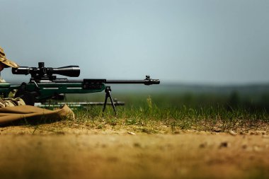 Bir keskin nişancı optik görüşlü bir tüfekle yere yatar. Kullanılmış mermi kovanı silahtan dışarı uçar.