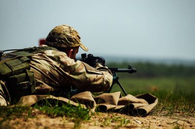 Bir keskin nişancı optik görüşlü bir tüfekle yere yatar. Kullanılmış mermi kovanı silahtan dışarı uçar.