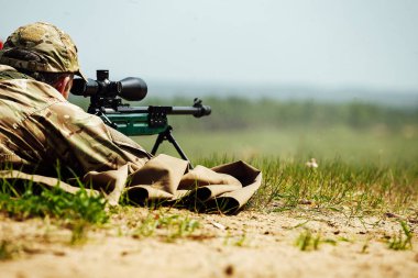 Bir keskin nişancı optik görüşlü bir tüfekle yere yatar. Kullanılmış mermi kovanı silahtan dışarı uçar.