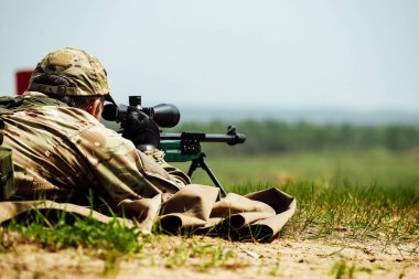 Bir keskin nişancı optik görüşlü bir tüfekle yere yatar. Kullanılmış mermi kovanı silahtan dışarı uçar.