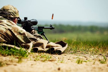 Bir keskin nişancı optik görüşlü bir tüfekle yere yatar. Kullanılmış mermi kovanı silahtan dışarı uçar.