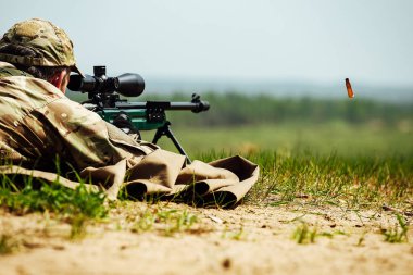 Bir keskin nişancı optik görüşlü bir tüfekle yere yatar. Kullanılmış mermi kovanı silahtan dışarı uçar.