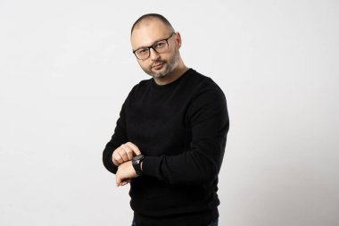 Kısa saçlı ve siyah çerçeveli gözlüklü genç bir adam kol saatinin beyaz bir arka planda durup kameraya baktığı zamanı gösteriyor.