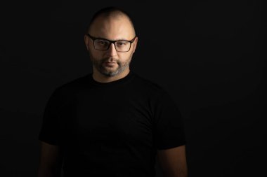 Siyah tişörtlü, kısa saçlı ve siyah çerçeveli gözlüklü genç bir adam karanlık bir arka plana dikkatlice ve agresif bir şekilde kameraya bakıyor.
