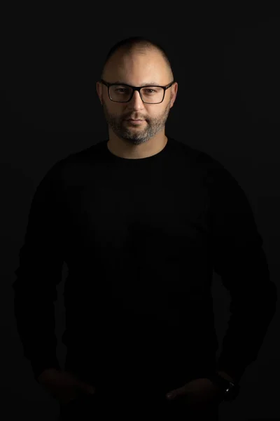 A young man in black-rimmed glasses and a black sweater keeps his hands in his pockets posing for the camera