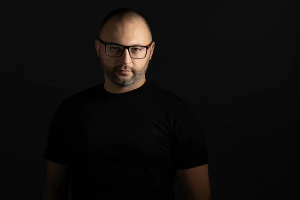 A young man in a black T-shirt with a short haircut and black-rimmed glasses poses against a dark background carefully and aggressively looking at the camera