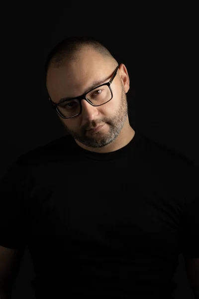 A young man in a black T-shirt with a short haircut and black-rimmed glasses poses against a dark background carefully and aggressively looking at the camera