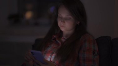 Gecenin geç saatlerinde evde yalnız bir kadın akıllı telefon ekranına bakıyor ve sıkılıyor.