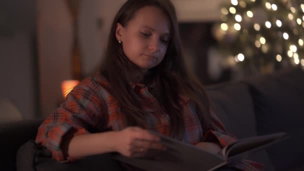 Uma mulher está sentada no sofá em seu apartamento e lendo um livro — Vídeo de Stock