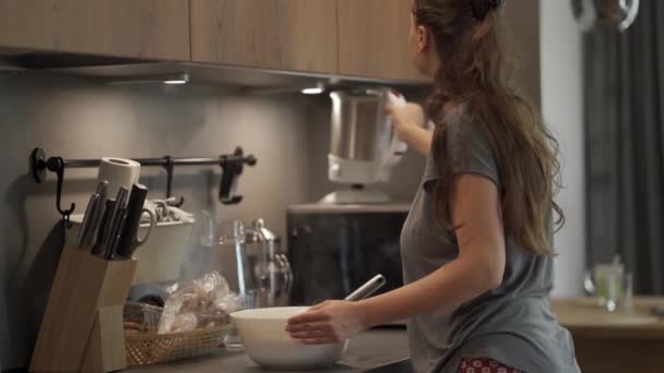 台所の自宅で長い髪の若い女性は 白いセラミックボウルに泡立て器で生地を攪拌することによって食べ物を準備します — ストック動画