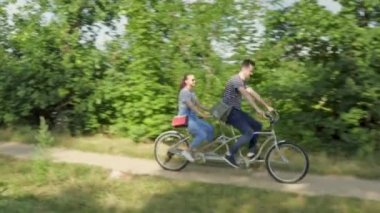 Genç evli bir çift, bir erkek ve bir kadın, nehir kıyısında yol boyunca iki koltuklu bir şehir bisikletine biniyorlar.