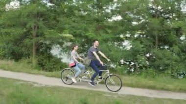 Genç evli bir çift, bir erkek ve bir kadın, nehir kıyısında yol boyunca iki koltuklu bir şehir bisikletine biniyorlar.
