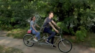 Genç evli bir çift, bir erkek ve bir kadın, nehir kıyısında yol boyunca iki koltuklu bir şehir bisikletine biniyorlar.