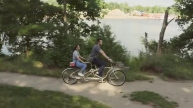 Genç evli bir çift, bir erkek ve bir kadın, nehir kıyısında yol boyunca iki koltuklu bir şehir bisikletine biniyorlar.