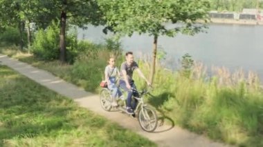 Genç evli bir çift, bir erkek ve bir kadın, nehir kıyısında yol boyunca iki koltuklu bir şehir bisikletine biniyorlar.