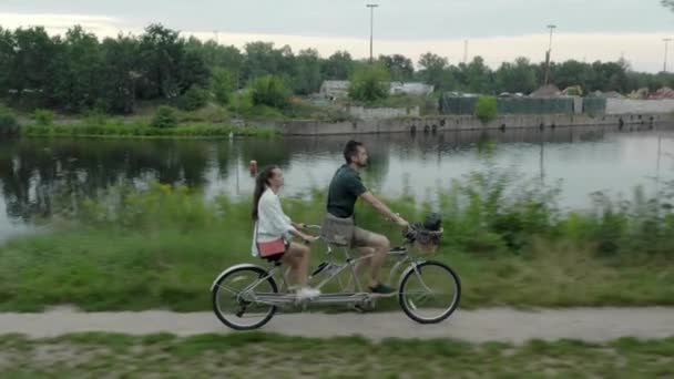 一对年轻夫妇 一个男人和一个女人 一起骑着一辆双座城市连载自行车沿着河边的公路骑着 — 图库视频影像