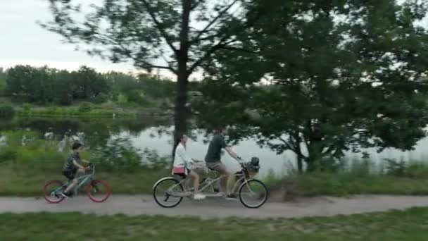 川沿いの道路に沿って2人乗りの都市タンデム自転車に乗る彼らの子供と一緒に若い夫婦 男性と女性は — ストック動画