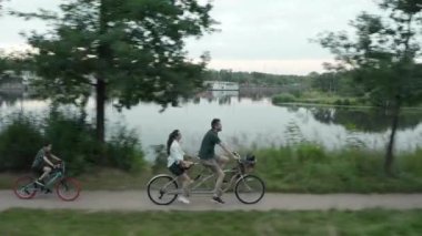 Genç evli bir çift, bir erkek ve bir kadın, çocuklarıyla birlikte nehir kıyısı boyunca iki koltuklu bir şehir bisikletine biniyorlar.
