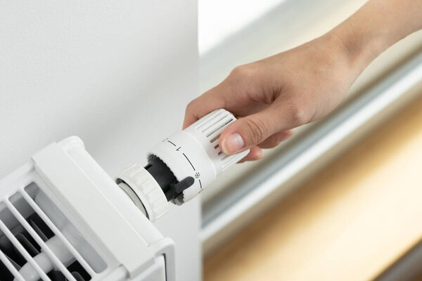 The woman turns off the heating by setting the regulator of the thermostatic valve of the radiator to the minimum value. Turning off the heating in the apartment during the energy crisis.