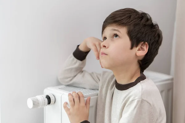 Çocuk ısınmak için ısıtıcının radyatörüne elleriyle sarılıyor. Soğuk kış mevsiminde enerji krizi sırasında konutların ısıtılması. Gaz ve elektrik kısıtlamaları ve tasarrufları.