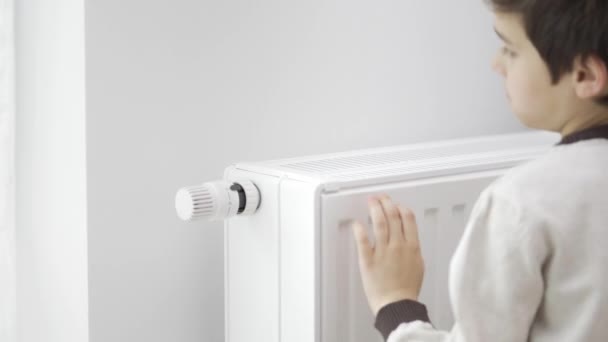 子供は冷凍手で家庭用ヒーターの冷たいラジエーターに触れ 暖めるために最大値で加熱力を増加させます その少年は暖房をつつく エネルギー危機時の暖房 — ストック動画