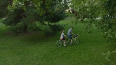 Genç evli bir çift, bir erkek ve bir kadın, güneşli bir yaz gününde iki kişilik bir bisikletle birlikte ağaçların arasındaki çimlerin üzerinde geziyorlar..