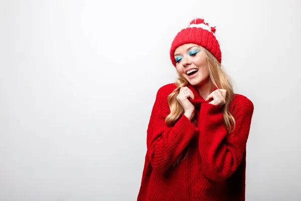 金发碧眼的金发女郎笑着 身穿红色毛衣 头戴一顶帽子 灰色背景上挂着明亮的冬装 — 图库照片