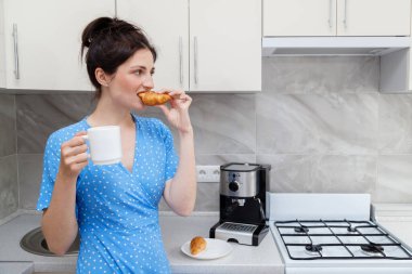 Bir kadın sabah kahve makinesinin yanında, mutfakta kahveli kruvasan yiyor. Kahvaltı.