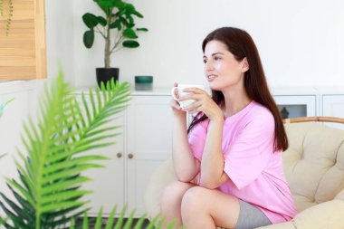 Oturma odasında beyaz bardaklı bir kadın içer ve pencereden dışarı bakar.