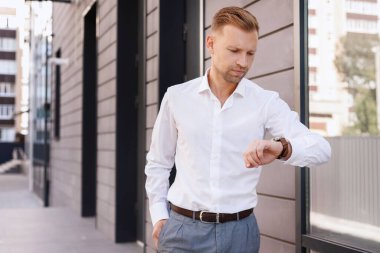 Ofis elbiseli çekici bir adam iş merkezinin yanındaki saatine bakıyor.