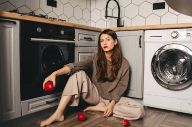 Kız mutfağının zemininde oturuyor. Kırmızı dudaklar ve kırmızı elmalar. Kırmızı elmalı parlak bir kız mutfağında yerde oturuyor..