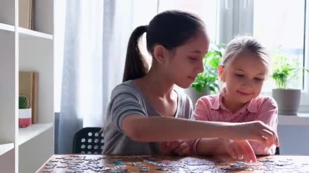 Dos hermanas-niñas lindas están felices de hacer rompecabezas, sentados en una mesa en la habitación. El concepto de infancia, juegos y ocio — Vídeo de stock
