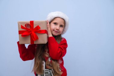Kırmızı Noel Baba şapkalı uzun saçlı kız Noel hediyesi olan bir kutu tutuyor ve yılbaşı hediyesini kulağının yanında tutuyor. Reklam için yer