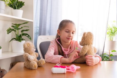 Pembe tişört giyen anaokulu öğrencisi bir kız hastanede oyuncaklarla oynuyor ve termometre oyuncak bir oyuncak ayı takıyor..
