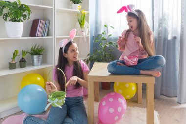 Pembe tişört ve tavşan kulağı giymiş iki kız kardeş Paskalya bayramını balonlarla süslenmiş bir odada bir sepette yumurta topluyor..