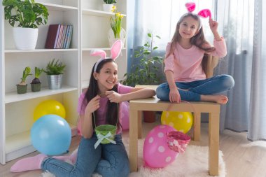 Pembe tişört ve tavşan kulağı giymiş iki kız kardeş Paskalya bayramını balonlarla süslenmiş bir odada kutlayarak bir sepette yumurta topluyor. Kameraya bakıyorlar..