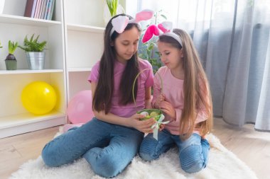 Pembe tişört ve tavşan kulağı giymiş iki kız kardeş Paskalya bayramını balonlarla süslenmiş bir odada bir sepette yumurta topluyor..