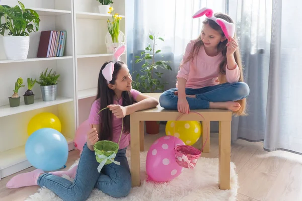 Dwie uśmiechnięte siostry ubrane w różowe t-shirty i królicze uszy zbierają jajka w koszyku, świętując Wielkanoc w pokoju ozdobionym balonami. — Zdjęcie stockowe