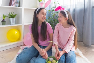 Pembe tişört ve tavşan kulağı giymiş iki kız kardeş Paskalya bayramını balonlarla süslenmiş bir odada bir sepette yumurta topluyor..