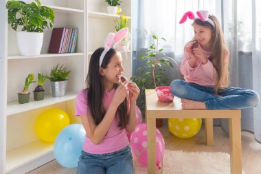 Pembe tişört ve tavşan kulağı giymiş gülümseyen iki kız kardeş bir sepette yumurta topluyor, balonlarla süslenmiş bir odada Paskalya bayramını kutluyorlar..