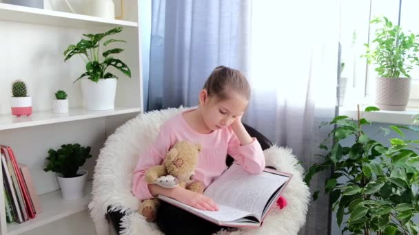 Ein trauriges Mädchen von 6-7 Jahren in einem rosa T-Shirt sitzt zu Hause auf einem Sessel und liest neben einem Bücherregal ein Buch und umarmt einen Teddybär — Stockvideo