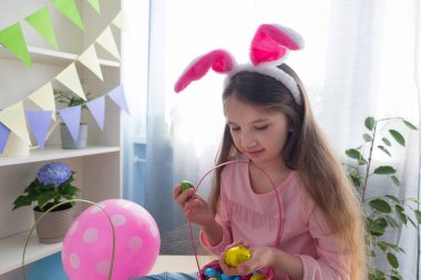 Pembe tişörtlü ve tavşan kulaklı bir kız Paskalya bayramını balonlarla süslenmiş bir odada kutluyor..