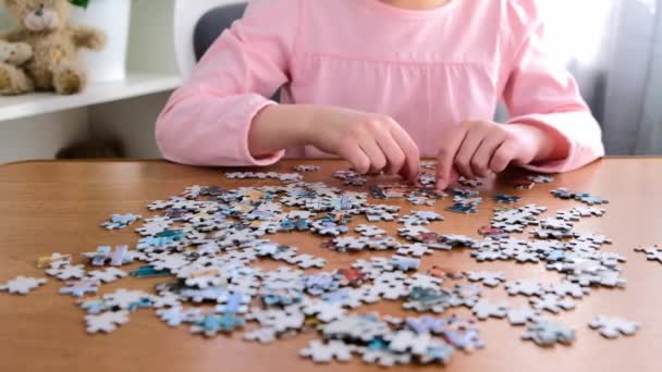 Bambina 6-7 anni in t-shirt rosa si siede a un tavolo a casa raccoglie puzzle. — Video Stock