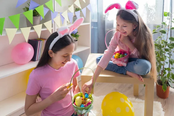 Pembe tişört ve tavşan kulağı giymiş iki kız kardeş Paskalya bayramını balonlarla süslenmiş bir odada bir sepette yumurta topluyor..